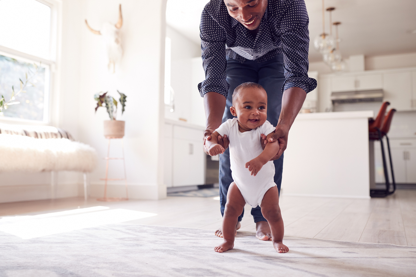 Father Encouraging Smiling Baby Daughter To Take First Steps And Walk At Home