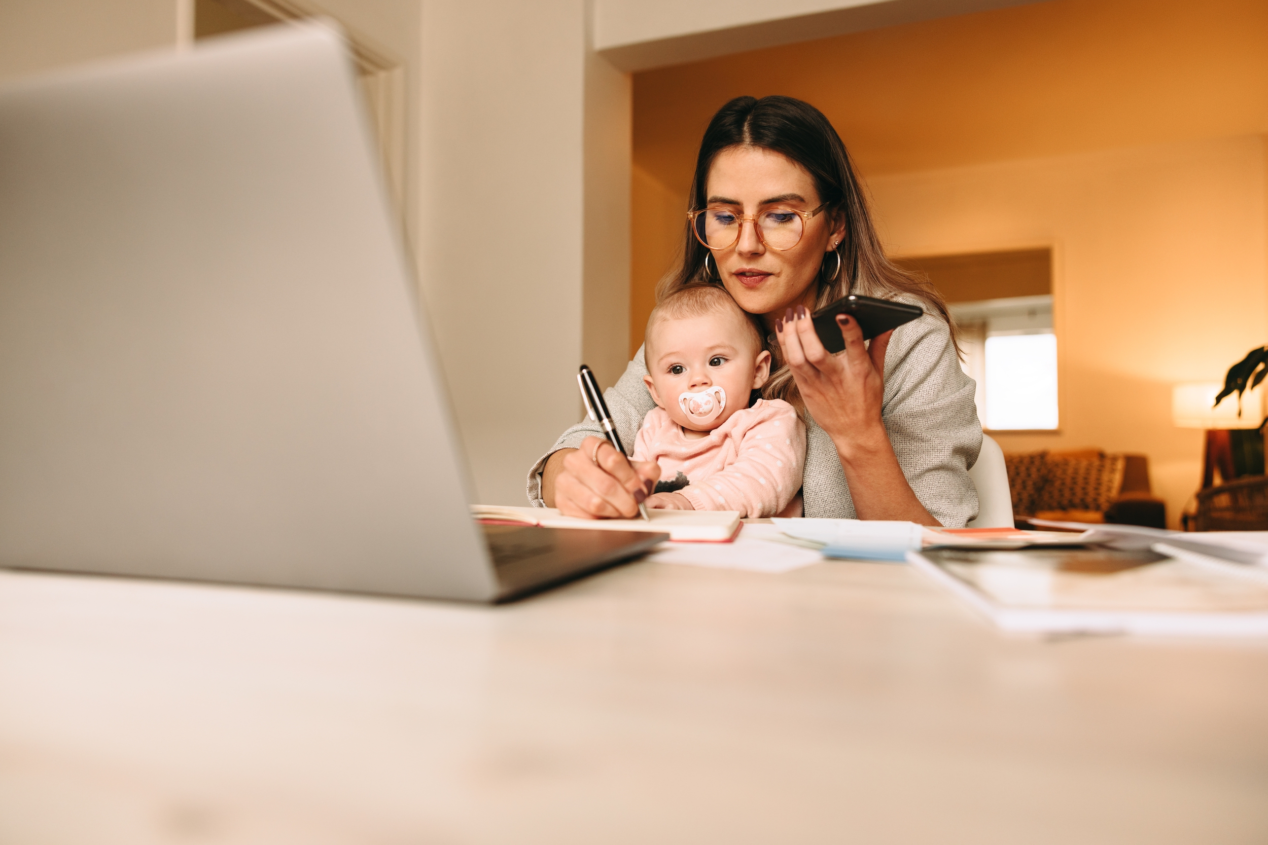 Working mom making notes during a phone call with her clients. Multitasking mom working on a new creative project in her home office. Female interior designer carrying her baby on her laps.